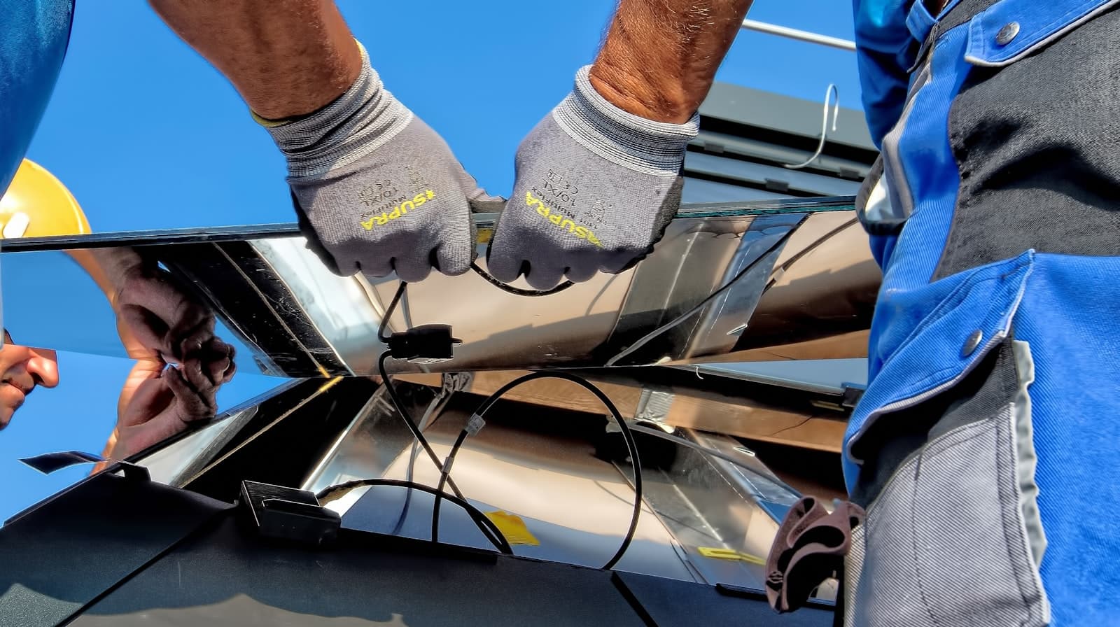 Gloved installer hands carefully fitting a solar panel rail on a roof under a blue sky